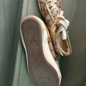 Bruna sneakers med guldiga detaljer från Coach - Snygga sneakers från Coach i brunt tyg med det klassiska monogrammönstret och guldiga paljett-detaljer på sidorna. Skorna har vita snören och vit sula med Coach-logga undertill. Perfekta för dig som vill sticka ut med lite extra bling.