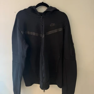 Svart hoodiejacka från Nike - Svart hoodiejacka från Nike med dragkedja framtill och huva. Jackan har diskret Nike-logga på bröstet och en stilren design med svarta detaljer. Nypris 1500.