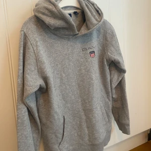 Grå hoodie från Gant - Säljer en grå hoodie från Gant med broderad logga och sköld på bröstet. Tröjan har huva och en stor magficka framtill. Perfekt för en avslappnad och stilren look. Liten fläck som bilden visar