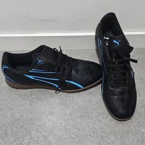 Svarta fotbollsskor från Puma med blå detaljer - Säljer ett par svarta fotbollsskor från Puma med blå detaljer och klassisk logga. Skorna har snörning och en platt sula med mönster för bra grepp. Perfekta för inomhusspel eller konstgräs.