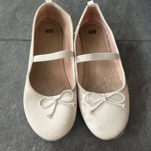 Vita ballerinaskor med rosett från H&M - Säljer ett par söta vita ballerinaskor från H&M med rund tå och dekorativ rosett framtill. Skorna har en elastisk rem över foten för extra bra passform. Perfekta för vår och sommar!