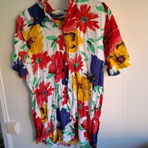Färgglad blommig skjortklänning - Säljer en skjortklänning med stora blommor i rött, gult och blått på vit botten. Klänningen har korta ärmar, krage och knäppning framtill. Perfekt för dig som gillar färg och mönster!