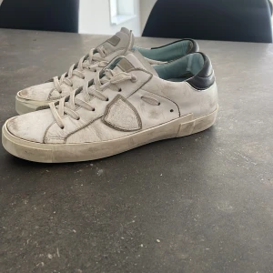 Vita sneakers från Philippe Model - Säljer ett par vita Philippe Model sneakers med svarta detaljer på hälen och gråa inslag vid sidan. Skorna har snörning och en platt sula. Perfekta för dig som gillar klassisk och stilren design.