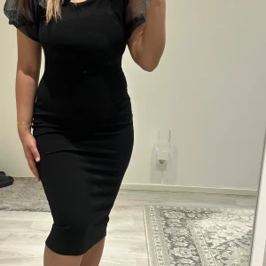 Svart tight klänning med puffärmar från Boohoo - Säljer en svart tight klänning från Boohoo med transparenta puffärmar och stor rosett på axeln. Klänningen är knälång och har en figurnära passform som framhäver siluetten.