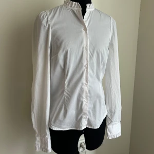 Vit skjorta med ståkrage och knappar - Säljer en klassisk vit skjorta med lång ärm och ståkrage. Blusen har knäppning framtill och fint avslut på ärmarna. Fin detalj under andra tröjor.