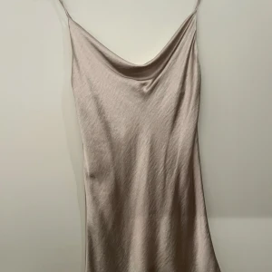 Champagnefärgad satinklänning med cowl neck - Säljer en champagnefärgad satinklänning med smala axelband och snygg cowl neck. Klänningen är kort och har en mjuk, glansig yta som ger en lyxig känsla. Perfekt för dig som vill ha en stilren och trendig look.