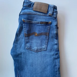 Blå jeans från Nudie Jeans - Säljer ett par klassiska blå jeans från Nudie Jeans med raka ben och snygga kontrastsömmar på bakfickorna. Jeansen har normal passform, knappgylf och är tillverkade i bomull. Perfekta för dig som gillar en tidlös stil.