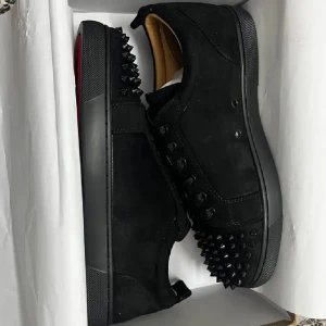 Louboutin skor - Säljer ett par svarta sneakers från Christian Louboutin med mattsvart ovandel i mocka och skinn, dekorerade med svarta nitar på tån och sidorna. Skorna har den ikoniska röda sulan och svarta skosnören.