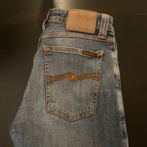 Blå jeans från Nudie Jeans - Säljer ett par klassiska blå jeans från Nudie Jeans med orangea sömmar och vågig brodyr på bakfickan. Jeansen har fem fickor och normal passform. Perfekta till vardags och lätta att matcha med olika stilar.