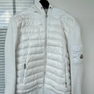 Vit pufferjacka/Cardigan med huva från Moncler - Säljer en stilren vit pufferjacka från Moncler med huva och dragkedja framtill. Jackan har quiltad framsida, två fickor med dragkedja och Moncler-logga på ärmen. Perfekt för kyligare dagar och riktigt snygg till streetwear. Jag har en även snören till de som kommer med 