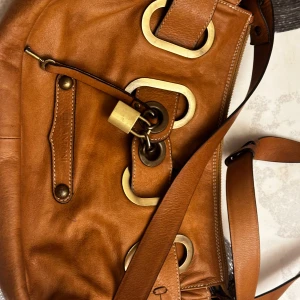 Brun axelväska från Antonia - Säljer en snygg brun axelväska från Antonia med stora guldfärgade metalldetaljer och justerbar axelrem. Väskan är i skinn och har ett dekorativt lås framtill samt innerfoder med logga. Perfekt för dig som vill ha en unik accessoar.