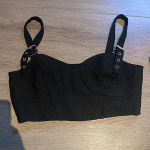 Svart croppad korsettopp från Divided - Säljer en svart croppad korsettopp från Divided med justerbara axelband med spännen. Toppen har en tight passform och elastisk rygg för extra komfort. Perfekt att styla till olika outfits!