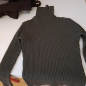 Mörkgrå ribbad polotröja - Säljer en mörkgrå ribbad polotröja med lång ärm. Tröjan har en hög polokrage och är stickad i ett mjukt material, perfekt för kyligare dagar. Enkel och stilren design som passar till det mesta.