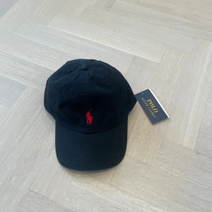Svart keps från Polo Ralph Lauren - Svart keps från Polo Ralph Lauren med röd broderad logga framtill och texten 'POLO' broderad baktill. Justerbart spänne bak för perfekt passform. Tillverkad i 100% bomull. Klassisk och stilren design. Classic Sports Cap Relay Black. Nypris 599 kr. Mitt pris 499 kr då den är helt ny med lapp dessutom. 