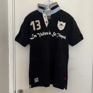 Svart pikétröja Les Voiles de St-Tropez - Snygg svart pikétröja med broderade detaljer, nummer 13 och text framtill. Märkeslogga på bröstet och ärmen, samt tryck och brodyr på ryggen. Krage i blåvitt med knappar och kontrastfärg. Perfekt för en sportig och stilren look.