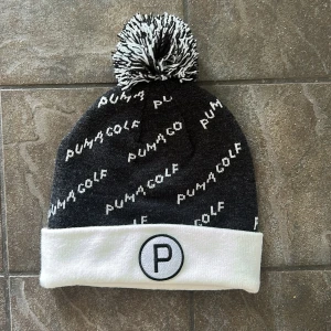 Svart och vit golfmössa från Puma - Snygg svart och vit mössa från Puma Golf. Mössan har Puma Golf-logga i vitt mönster över hela och ett broderat P framtill. Perfekt för kyliga dagar på golfbanan.