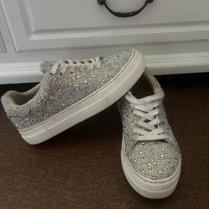 Glittriga sneakers med platåsula - Säljer ett par snygga sneakers från blue by betsey johnson täckta med glittrande stenar. Skorna har vit platåsula, rund tå och vita snören. Perfekta för dig som vill sticka ut med lite extra bling i vardagen. Köpte för 1350kr 