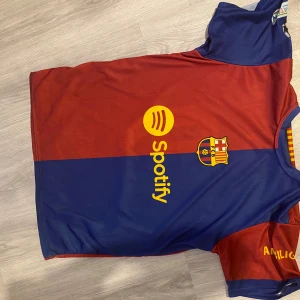 FC Barcelona matchtröja Lamine Yamal 19 - Säljer en officiell FC Barcelona fotbollströja med Lamine Yamal och nummer 19 på ryggen. Tröjan är röd och blå med klubbmärke, Spotify-logga och UNHCR-tryck. Kortärmad modell i lätt och ventilerande material, perfekt för match eller träning.
