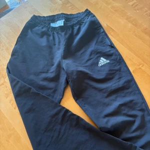 Adidas windbreaker byxor - Svarta träningsbyxor från Adidas med vit logga på benet. Storlek S och sparsamt använda. Väldigt sköna och passar för sommar, vår och höst väder. Köpta för 600. Tveka inte att höra av er vid frågor och intresse!