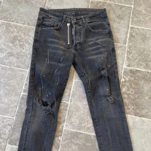 Svarta slitna jeans från Dsquared2 - Säljer ett par svarta jeans från Dsquared2 med slitningar och färgstänk på benen. Jeansen har dragkedja, fem fickor och klassisk Dsquared2-logga vid gylfen. Snygg, smal passform och cool streetstil.