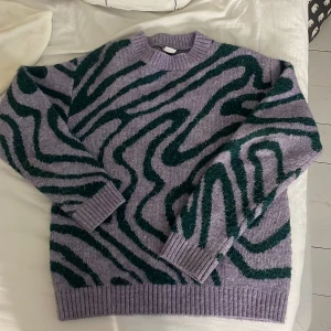 Lila och grön mönstrad stickad tröja från Lindex - Säljer en stickad tröja från Lindex i lila med grönt abstrakt mönster. Tröjan har rund halsringning, långa ärmar och breda ribbade muddar vid ärmslut och nederkant. Perfekt för dig som gillar färg och mönster!