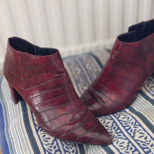 Vinröda boots med krokodilmönster - Säljer ett par vinröda boots  str 37 med krokodilpräglat mönster och spetsig tå. Skorna har klack och dragkedja på insidan för enkel påtagning. Bekvämklack, bekväm i sida  snygga och unika  nyskick