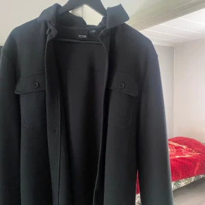 Svart overshirt med knappar - Stilren svart overshirt med klassisk krage och två bröstfickor med lock och knappar. Jackan har långa ärmar och stängs med knappar framtill. Perfekt för lager-på-lager och enkel att matcha med olika outfits.