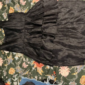 Svart volangklänning från H&M - Säljer en svart klänning från H&M med volangdetaljer och ärmlös design. Klänningen har en elegant struktur i tyget och markerad midja. Perfekt för dig som vill ha en stilren och trendig look.