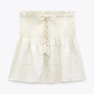 Vit kjol med smock och knytband - Supersöt vit kjol med smock upptill och dekorativt knytband framtill. Kjolen har en lätt volang och är perfekt för varma dagar. Den är kort och har en luftig känsla som passar till många olika stilar.