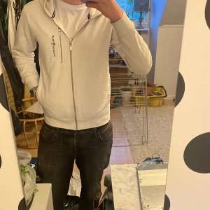 Vit hoodie från Sail Racing - Säljer en stilren vit hoodie från Sail Racing med dragkedja framtill och på bröstfickan. Jackan har broderad logga på bröstet och huva. Perfekt för en avslappnad look. Några små skador som inte syns alls när du har på dig den(fläckar) därav billig!!!!