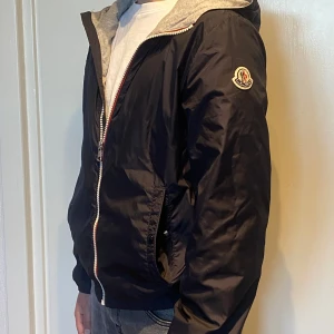 Marinblå vindjacka från Moncler.  - Säljer en marinblå vindjacka från Moncler med grå huva och dragkedja framtill. Jackan har Moncler-logga på ärmen och innerfoder i grått. Perfekt för dig som vill ha en stilren och trendig jacka. Säljer för att den är för liten! Mycket bra skick! Skanna QR-koden för bevis på att den är äkta. 😊 storleken är 12A - 12 år vilket är cirka 150-152. 