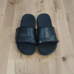 Svarta sandaler i syntetmaterial - Svarta sandaler med bred rem över foten och platt sula. Tillverkade i syntetmaterial med enkel och stilren design. Perfekta för varma dagar eller att ha hemma.