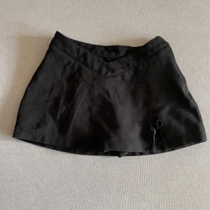 Svart kortkjol med shorts under - Säljer en svart kortkjol med inbyggda shorts och dragkedja i sidan. Kjolen har en enkel design och är perfekt för dig som vill ha både stil och komfort. Passar till många olika tillfällen och är lätt att matcha med olika toppar.