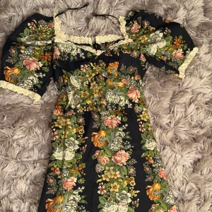 Blommig långklänning med puffärmar och spetsdetaljer - Superfin långklänning med svart botten och färgglatt blommönster i grönt, rosa, gult och vitt. Klänningen har puffärmar, spetskant vid halsringning och ärmslut samt volang nertill. Perfekt för dig som gillar romantisk och vintageinspirerad stil.