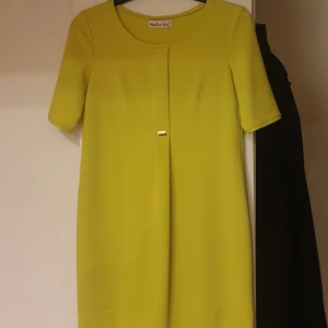 Gul klänning med rosettdetalj från Michael Kors - Snygg gul / limegrön klänning med korta ärmar och en liten svart rosett baktill vid halsen. Klänningen har rak passform och är perfekt för dig som vill sticka ut med färg. Enkel och stilren design.