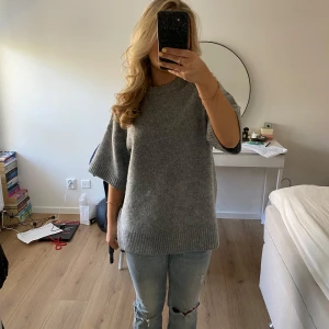 Grå stickad tröja med kort ärm - Mysig grå stickad tröja med korta, vida ärmar och rund hals. Perfekt att slänga på sig över en t-shirt eller långärmad topp. Enkel och stilren design som passar till det mesta.