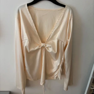 Beige satinblus med knyt framtill - Säljer en beige långärmad blus i satin med djup v-ringning och knyt framtill. Super att ha till fest, aldrig använd😊