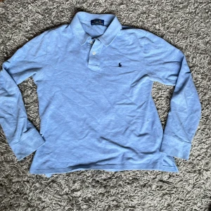 Ljusblå långärmad pikétröja från Polo Ralph Lauren - Säljer en klassisk ljusblå långärmad pikétröja från Polo Ralph Lauren. Tröjan har krage, knappar vid halsen och den ikoniska broderade loggan på bröstet. Perfekt för en stilren och avslappnad look. Hör av dig om frågor.