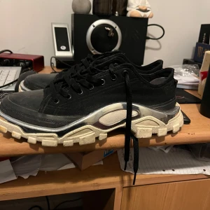 Adidas x Raf Simons Detroit Runner svarta sneakers - Säljer ett par svarta Adidas x Raf Simons detroit runner sneakers med vit och silverfärgad sula. Skorna har snörning och en unik futuristisk design med blandade material och detaljer. Perfekta för dig som gillar statement-sneakers.