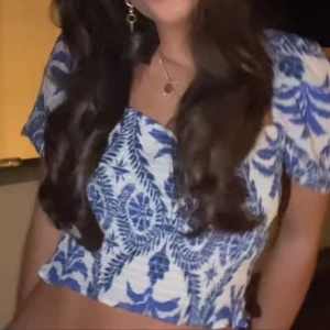 Blåvit blus med puffärmar - Säljer en blåvit blus med tropiskt mönster och puffiga korta ärmar. Toppen är croppad och har en figurnära passform. Perfekt för sommardagar och ger en fräsch look till jeans eller kjol.