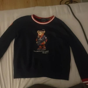 Mörkblå tröja med Polo Bear från Ralph Lauren - Säljer en mörkblå långärmad tröja från Ralph Lauren med Polo Bear-motiv på bröstet och USA Olympic Team-märke på ärmen. Röd och vit rand vid kragen och ärmslut. Perfekt för dig som gillar sportig stil.