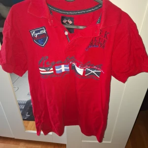 Röd pikétröja från J.F.K med tryck - Röd pikétröja från J.F.K med broderade och tryckta detaljer, bland annat flaggor och texten 'Regatta Sailing' samt 'Blue Water'. Tröjan har korta ärmar, krage och knappslå framtill. Perfekt för en sportig look.