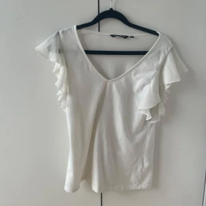 Vit topp med volangärmar från KappAhl - Säljer en vit topp från KappAhl med v-ringning och volangdetaljer på ärmarna. Toppen har en feminin och luftig design, perfekt för varmare dagar. Enkel att matcha med både jeans och kjol.