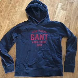 Mörkblå hoodie från GANT - Säljer en mörkblå hoodie från GANT med röd text 'NEW HAVEN GANT EAST COAST 1949' på bröstet. Tröjan har huva, magficka och ribbade muddar vid ärmslut och nederkant. 