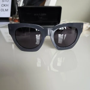 solglasögon från Ace & Tate - Snygga solglasögon i en cool grå färg från Ace & Tate. Modellen har en markerad, kantig båge med breda skalmar och mörka glas. 