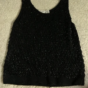 Svart pärlbroderad linnetopp från Intuitions - Säljer en svart linnetopp från Intuitions i 100% ull, dekorerad med svarta pärlor över hela framsidan. Toppen är ärmlös och har en klassisk rund halsringning. Perfekt för att lyfta en outfit med lite extra detaljer.