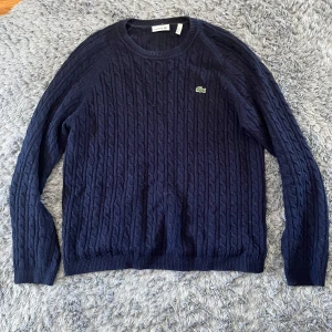 Mörkblå stickad tröja från Lacoste - Säljer en mörkblå stickad tröja från Lacoste med klassiskt kabelstickat mönster och rund halsringning. Tröjan har lång ärm och den ikoniska gröna krokodilloggan på bröstet. Perfekt för kyliga dagar.