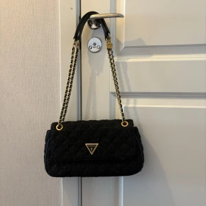 Väska från Guess - Svart, glitttig väska från Guess med guldiga detaljer och kedja, använd ett fåtal gånger. Pris kan diskuteras 