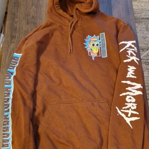 Brun Rick and Morty hoodie - Säljer en brun hoodie med Rick and Morty-tryck på bröstet, ryggen och ärmarna. Tröjan har huva med dragsko och en stor magficka. Färgglada detaljer och texten 'WUBALUBBADUBDUB' på ärmen.