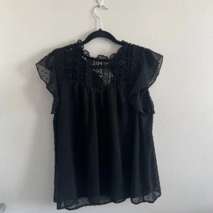 Svart blus med spets och volang - Säljer en svart blus med v-ringning, spetsdetaljer upptill och volang vid ärmarna. Blusen är ärmlös med lätt transparent tyg och små prickar i materialet. Perfekt för dig som gillar romantiska detaljer.